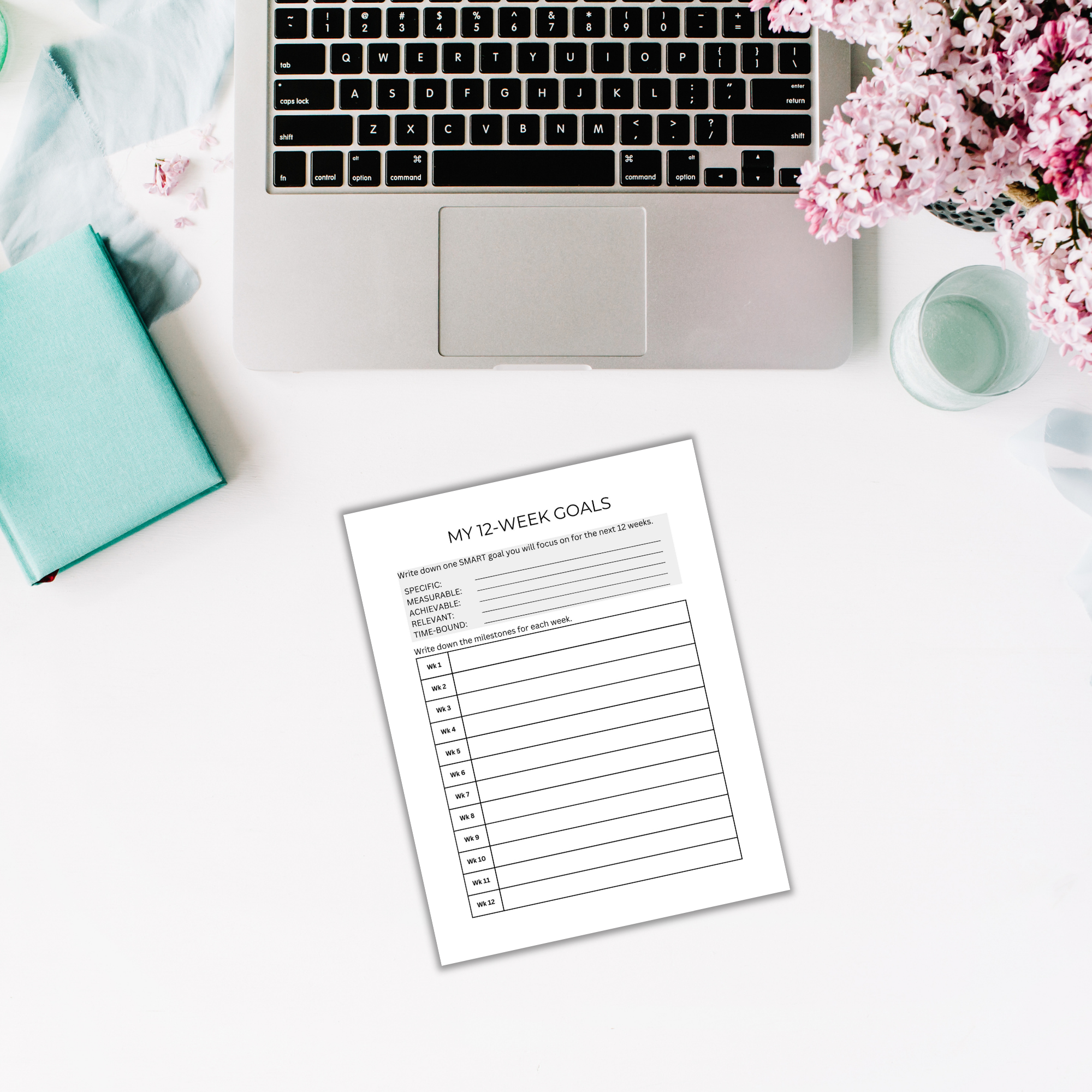 A laptop on a white desk sits alongside a 12-Week Success Sprint Planner PLR Template, a teal notebook, a pen, and a vase of pink flowers.