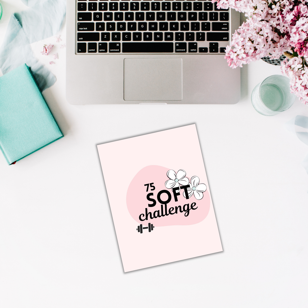 A laptop, open notebook, and vibrant flowers are neatly arranged on a white table alongside 'The 75 Soft Challenge' Trackers booklet with handy organizers.