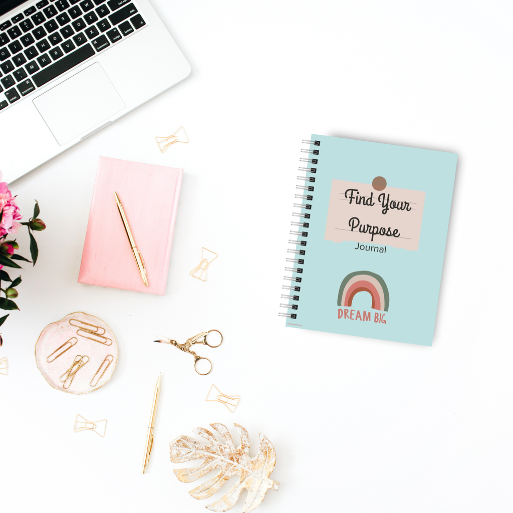 A laptop, pink notebook, and The 'Find Your Purpose' Journal are arranged on a white desk decorated with pink flowers, gold paperclips, and scissors.