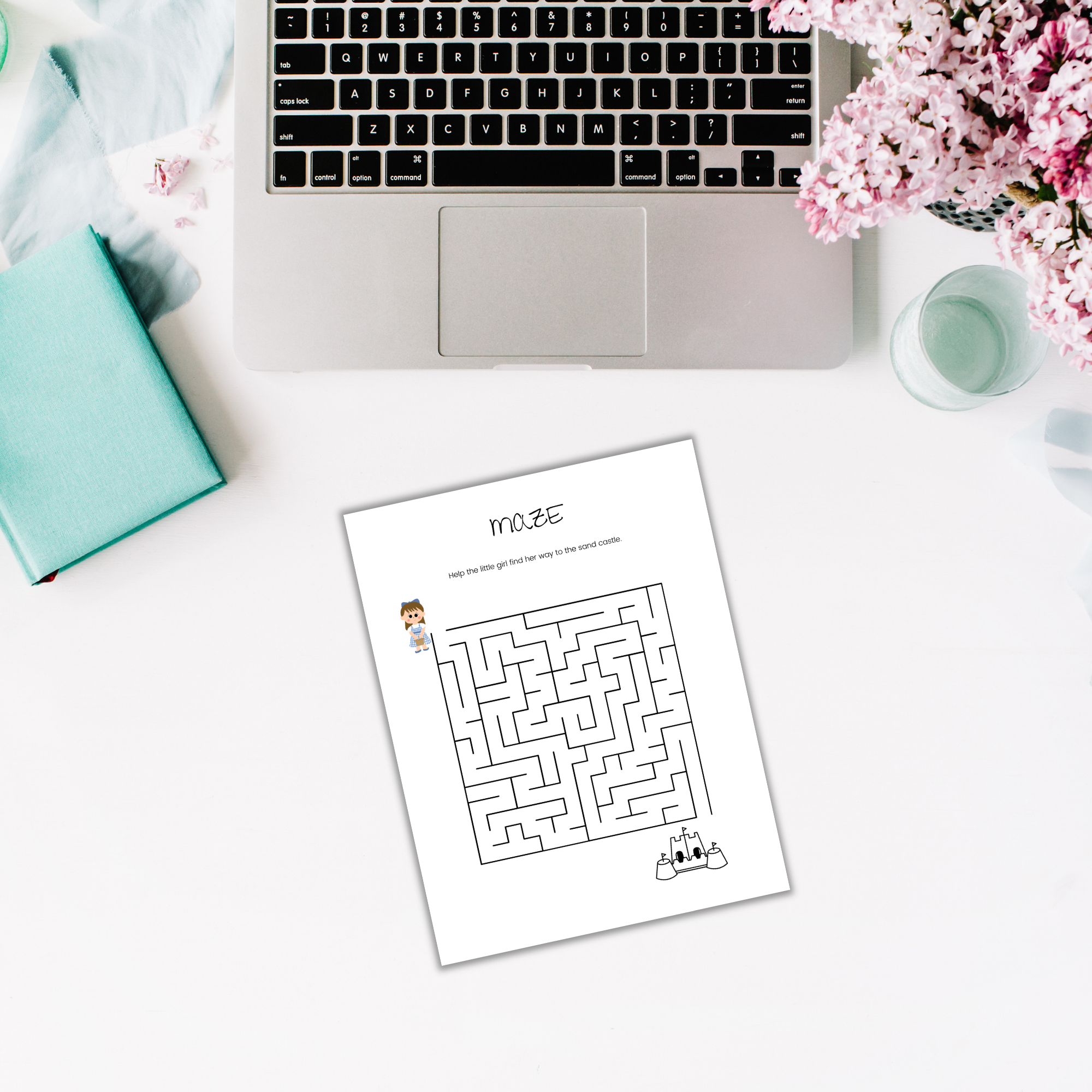 A laptop, an open notebook, a glass, a bouquet of pink flowers, and a printed maze from the Summer Activity Pack for Kids rest on the white desk.