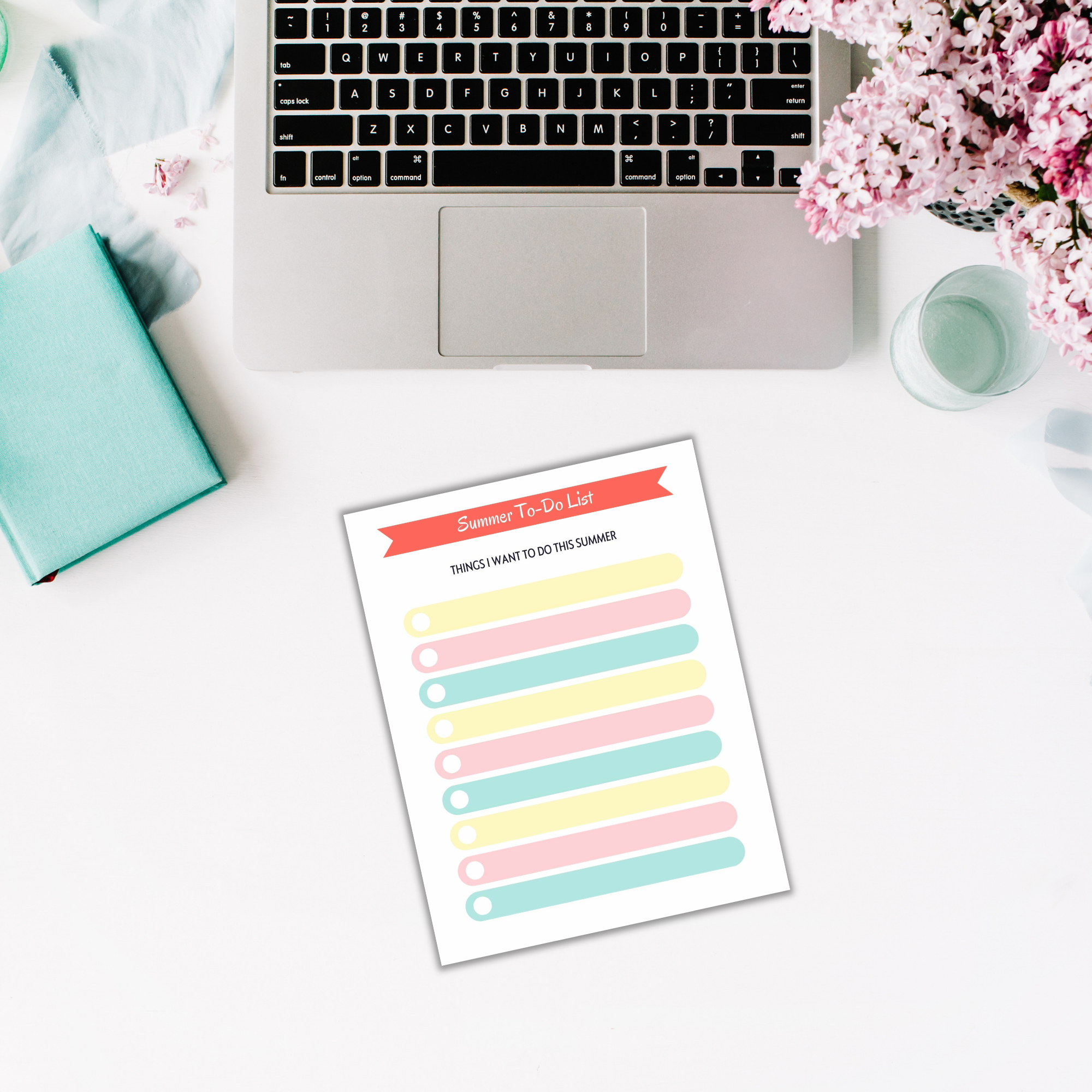 A laptop, notebook, and a Summer Planner for Kids filled with vibrant activities adorn a white desk decorated with pink flowers.